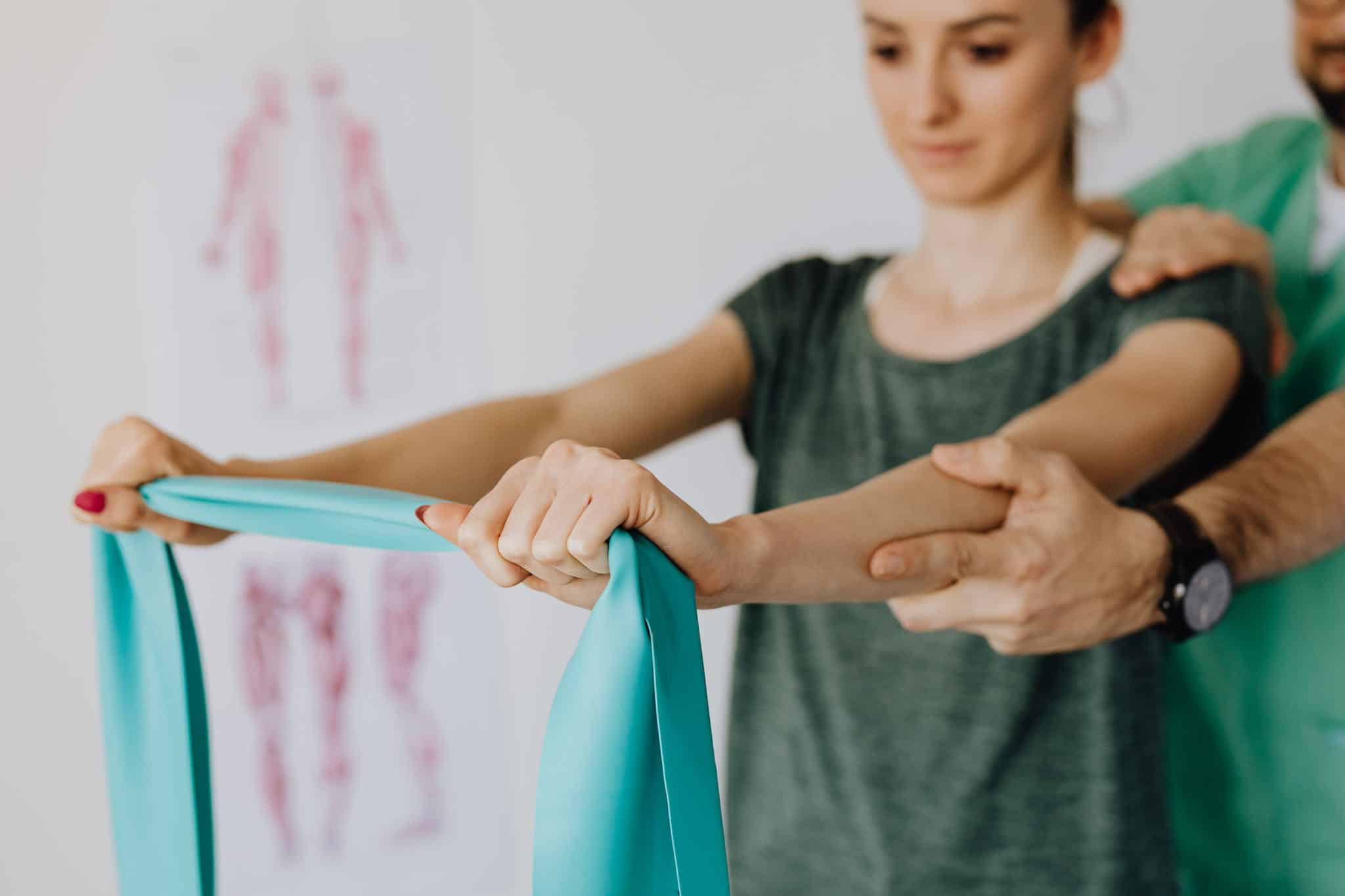 Crop anonymous orthopedist in wristwatch and uniform helping young woman in casual wear reaching arms with elastic tape in doctor office